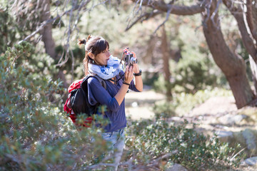 Fototapeta premium Mid adult backpacker with ponytail standing in th forest while using a photography camera to take a shoot.