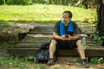 Asian man in black casual wear wearing a flu mask sitting on old boards of a wooden bridge in public park with grreen lawn nature background