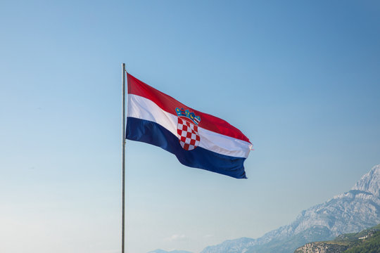 The red white blue Croatian flag waving in the wind on a flagpole on a sunny day and a blue sky, with in the background the Biokovo mountains on Makarska, Dalmatia at the Mediterranean coast