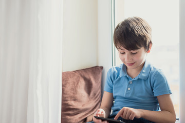 Boy playing with tablet sitting on pillow and windowsill. Soft focus. Quarantine and online school concept