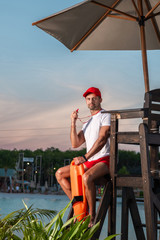 Portrait of a beautiful lifeguard with a whistle in his hands, sitting on a rescue tower and posing...