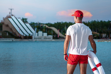 Rear view: A male lifeguard on the beach holds a lifebuoy in his hands and looks into the distance. Blurred background of water park