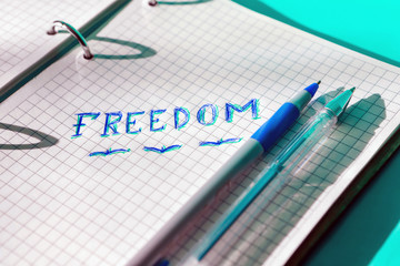 The inscription freedom in an open notebook in a cage. Written in blue and green pens. The pens are on the notebook near the inscription.