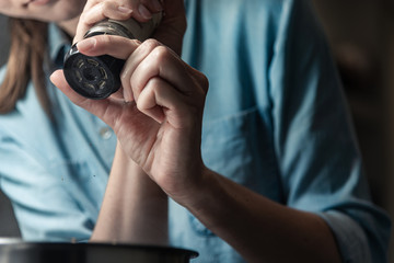 Woman adding black pepper with grinder to the cooking food in a saucepan.