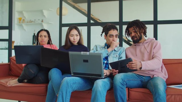 Angry frustrated young asian woman caught her arabic boyfriend watching embarrassing content online on digital tablet with male friend, smacking him upside the head while sitting on sofa at home.