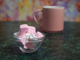 a bowl with pieces of white and pink marshmallows is on the green table. pink coffee cup on the background. close up. copy space