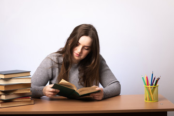 a young girl sits at a table and reads a book. student preparing for the exam. gray wall on the background. a jar with multi-colored pencils stands nearby.