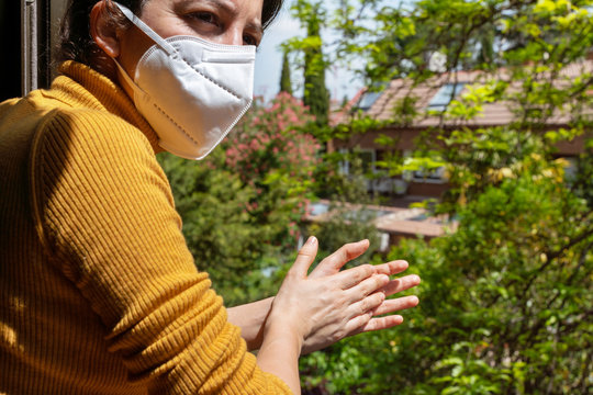 Unrecognizable Woman Wearing A Mask Clapping To Medical Community Through The Window. Covid 19 Coronavirus Obligation To Stay Home During Pandemic Disease. People Lifestyles Quarantine In The House.