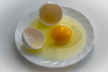 One cracked egg with yellow yolk surrounded by egg shells is laying on a white plate on white background
