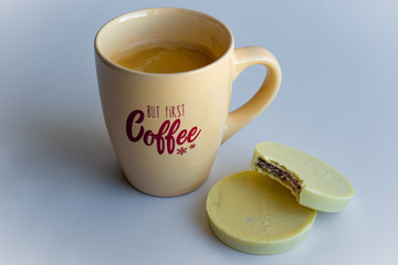 View of a coffee cup with lettering full with black coffee together with round cookies enrobed with white chocolate on white background