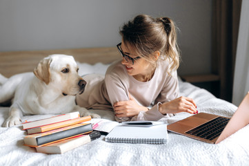 An attractive young girl is learning online using books and a video lesson on a laptop. Next to her...