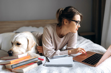 Fototapeta na wymiar An attractive young girl is learning online using books and a video lesson on a laptop. Next to her on the bed lies her cute dog. Learning at home