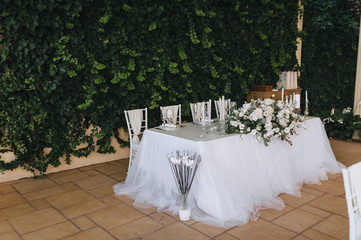 A wedding table decorated with flowers, dishes, candles, glasses, chairs stands against a background of blooming green ivy. Ceremony, party. Photography, concept.