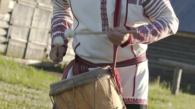 Man in traditional russian clothes dancing and playing on folklore drum outdoor on traditional antique wooden windmill background. Man wearing national Finno-Ugric clothes.