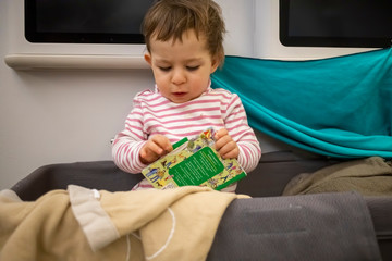 little cute toddler sitting in the baby bassinet of an airplane sitting and leafing through a...