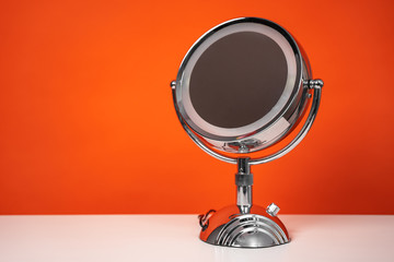 Round mirror on metal stand with backlight stands on table on orange background, copy space for text, front view. Beauty equipment for makeup artists and cosmetologists for use at home.