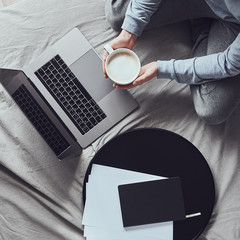 Young woman wearing gray home clothes sits on a bed in front of laptop and holds a mug of...