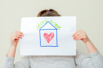 Closeup of child's hands holding a picture of red house with heart and words Stay Home covering her face. Stay Home concept.