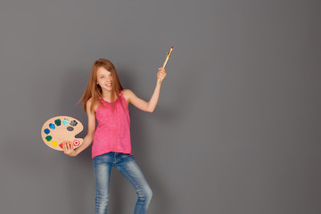 A girl with a palette and brushes, laughing against a gray wall. Wants to paint it with bright colors. The concept of children's creativity. Space for your text