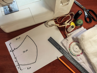 Pattern of medical face mask drawn on a white paper on the background of sewing accessories. Sewing machine, white cloth, scissors, ruler, pencil, thread and other materials on the brown surface