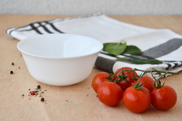 White bowl and a sprig of cherry tomatoes on a wooden table, green leaves and kitchen towel are visible in the background