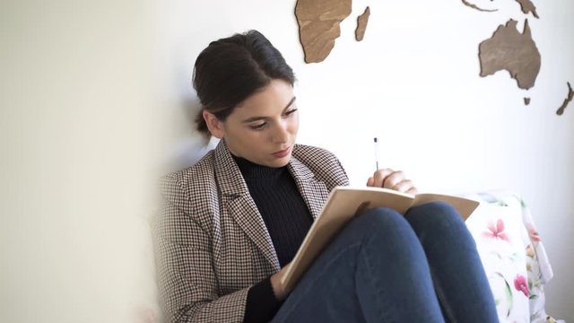 Chica joven atractiva escribe en una libreta sentada en la cama de su habitaci&oacute;n.