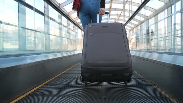 Unrecognizable young girl walking on escalator with her luggage. Legs of sexy woman going and pulling suitcase on wheels. Lady going to trip. Concept of travel or holiday. Slow motion Back view