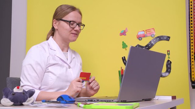 Young speech therapist, woman in white coat, providing the online lesson using laptop, showing how to pronounce the words, strongly articulating. The session for child, with children pictures around