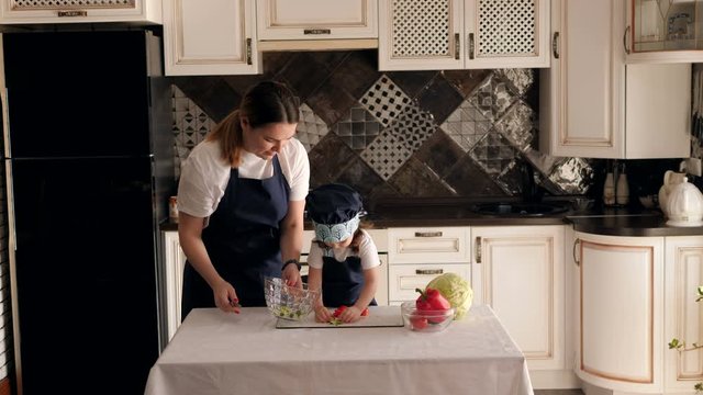 Close-up of a little girl helping her mother prepare a vegetable salad, she puts sliced cucumber in a plate. Slow motion. Little baby cook.