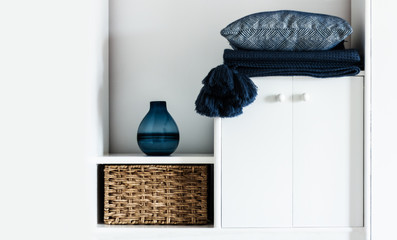 Cozy home interior decor: pillow, plaid, blue vase,  wicker box on a white shelf in the room. The quarantine concept of stay home, distance home education background.