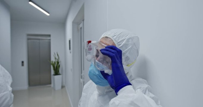 Tired female medical worker with face scars from protective uniform after overwork with patients coronavirus infected taking off protective mask and resting in corridor of modern clinic