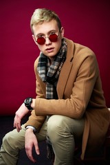 Handsome young man in stylish sunglasses and scarf posing against red studio background 