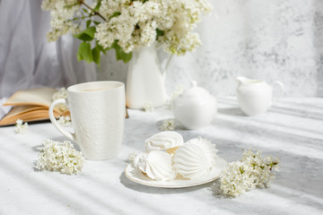 Still life vase with a bouquet of white lilac, a Cup of coffee with marshmallows, a plate of meringue, a sugar bowl and a milk jug , an old book. Postcard good morning.