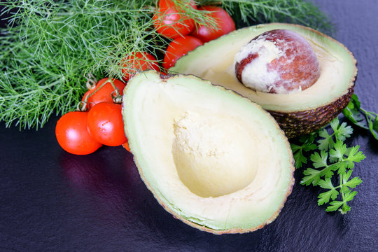 Halves of fresh big avicado with tomatoes cherry and heap of dill and parsley on the black background. Ketogenic diet food avocado.