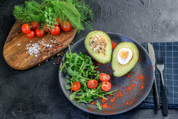 Halves of fresh avocado filling with eggs and tuna with sauce, tomatoes cherry and green fresh condiments dill, parsley and arugula. Black dish, black cutlery and napkin on the black background.