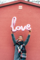 Pleasant young woman in a coat near the pink wall holds the inflated word LOVE