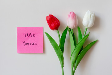 Text love yourself on short note paper with tulips flowers on white background.