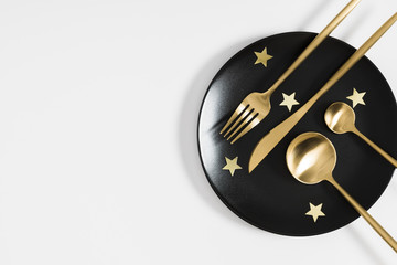 Black empty ceramic plate and golden cutlery, golden stars on white background. Holiday dinner concept. Flat lay, top view, copy space