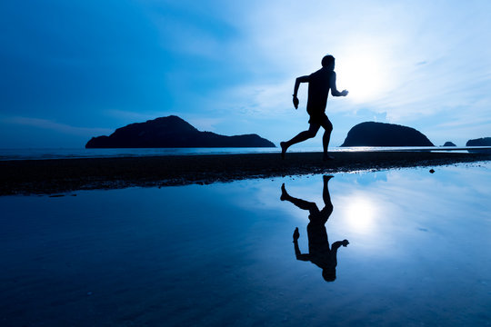 Silhouette Asian man running on the beach with shadow on water during sunrise toned in blue color