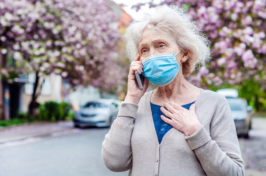 An old woman in a protective mask on her face is talking on her cell phone walking in the spring flowering town. COVID-2019, coronavirus. Senior Health. Weakening quarantine. Outdoors walks