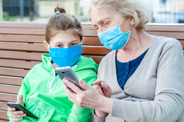 The child helps grandmother. Grandmother with a grandson in protective medical masks are sitting on a bench with mobile phones. Epidemic coronavirus covid-19. Walking outdoors. Family relationships