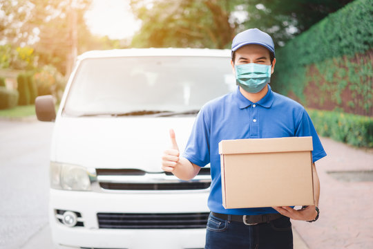 Postal delivery courier man thumbs up wearing protective face mask in front of cargo van delivering package holding box due to Coronavirus disease or COVID-19 outbreak situation in the world.