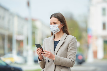 A girl with long hair in a medical face mask to avoid the spread coronavirus uses a smartphone in the street. A woman in a face mask against COVID-19 wears a coat waits in the center of the city.