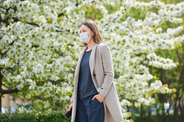 A girl in a medical face mask to avoid the spread coronavirus holds a smartphone in the park. A woman in a face mask against COVID-19 keeps a social distance in the garden between flowering trees.