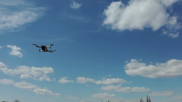 A small gray drone flying over the city against a bright blue sky with fluffy clouds. new delivery technologies and video photos. drone in flight over the houses and high-rise buildings of the city