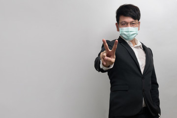 Asian businessman wearing surgical face mask in formal black suit jacket, raise 2 fingers up, victory sign by right hand, look at the camera, isolate with grey background, concept coronavirus, covid19