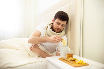 Fototapeta na wymiar Sick Man in Bed Adding Lemon into Tee. Man Ill in Bed Self Healing. Self Treatment at Home. Man Drinking Hot Tea in Bed.