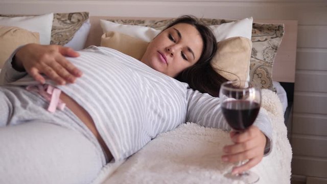 Sad pregnant woman lying on the bed with a glass of wine. Unwanted pregnancy. A pregnant girl drinks alcohol.