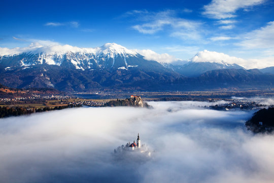 amazing panorama of Lake Bled Blejsko Jezero on a foggy morning with the Pilgrimage Church of the Assumption of Maria on a small island and Bled Castle and Julian Alps in backgroud © Melinda Nagy