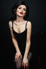 Gorgeous young brunette woman in elegant black dress posing against dark studio background. Sensual young woman with red lips and bright makeup.